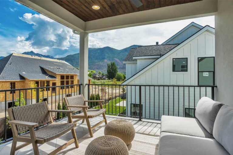 The image shows a balcony with two chairs and an ottoman overlooking mountains from a house.