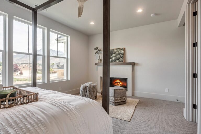 A room with a bed and fireplace has white walls and windows showing an outdoor view.