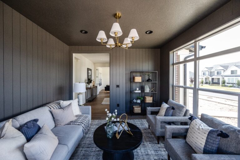 A room with grey walls and a chandelier has white furniture and large windows showing outside houses.