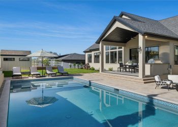 A house with a pool and chairs is surrounded by trees under clear skies.